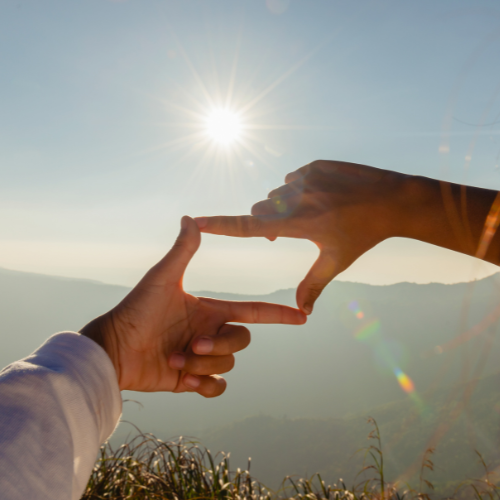 Zwei Hände formen einen rechteckigen Rahmen vor einer sonnigen Berglandschaft.